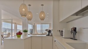 Une cuisine moderne avec des meubles blancs, un grand îlot central, des luminaires suspendus et une vue sur la mer.