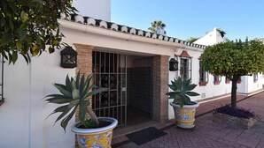 Un bâtiment à la façade blanche, doté d’un toit en tuiles et d’un portail en métal. Deux plantes en pot sont placées devant l’entrée.