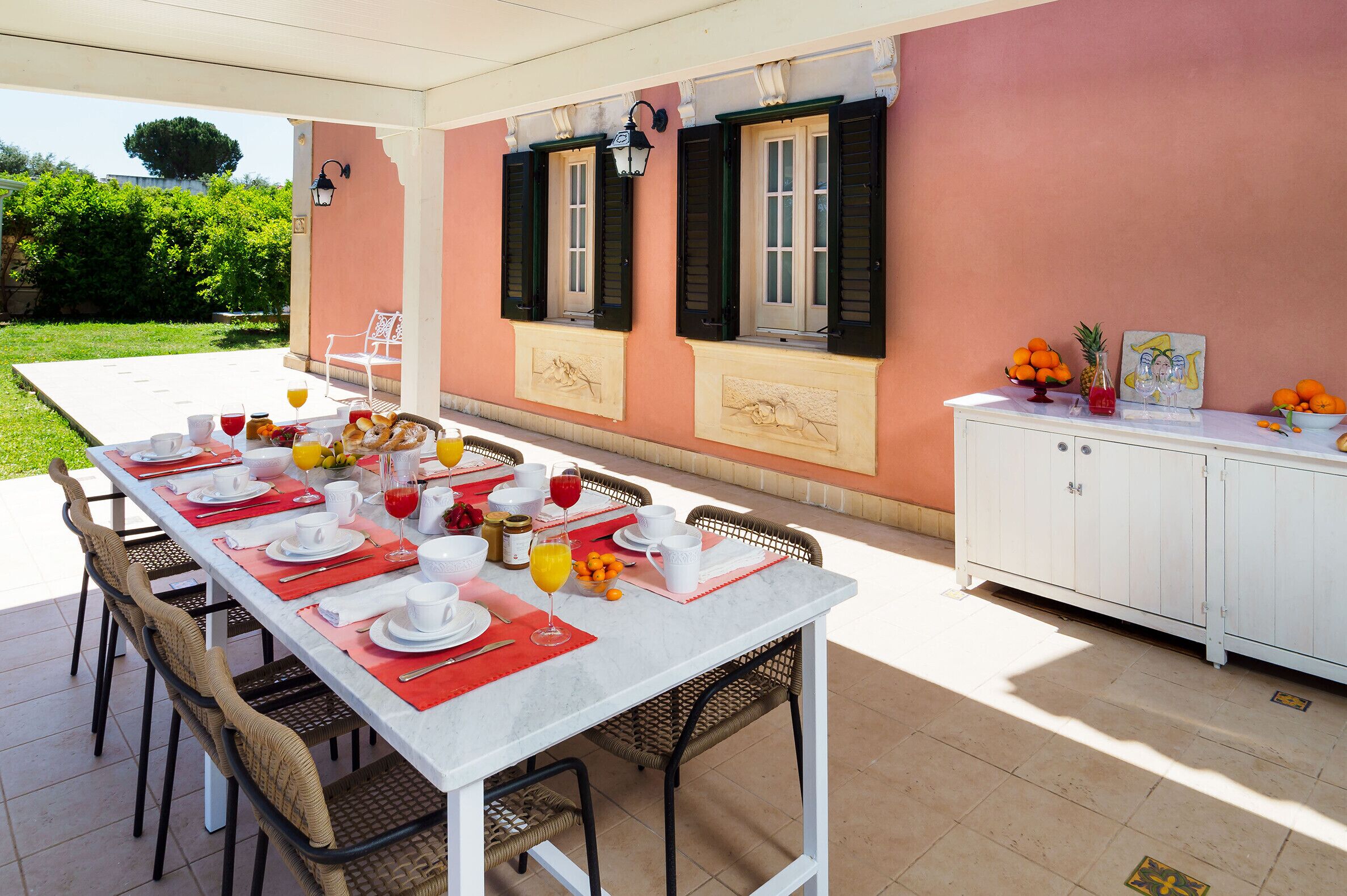 An outdoor dining area with a table set for a meal, surrounded by chairs, and a white cabinet with fruit and a painting on top.