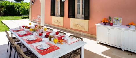 An outdoor dining area with a table set for a meal, surrounded by chairs, and a white cabinet with fruit and a painting on top.