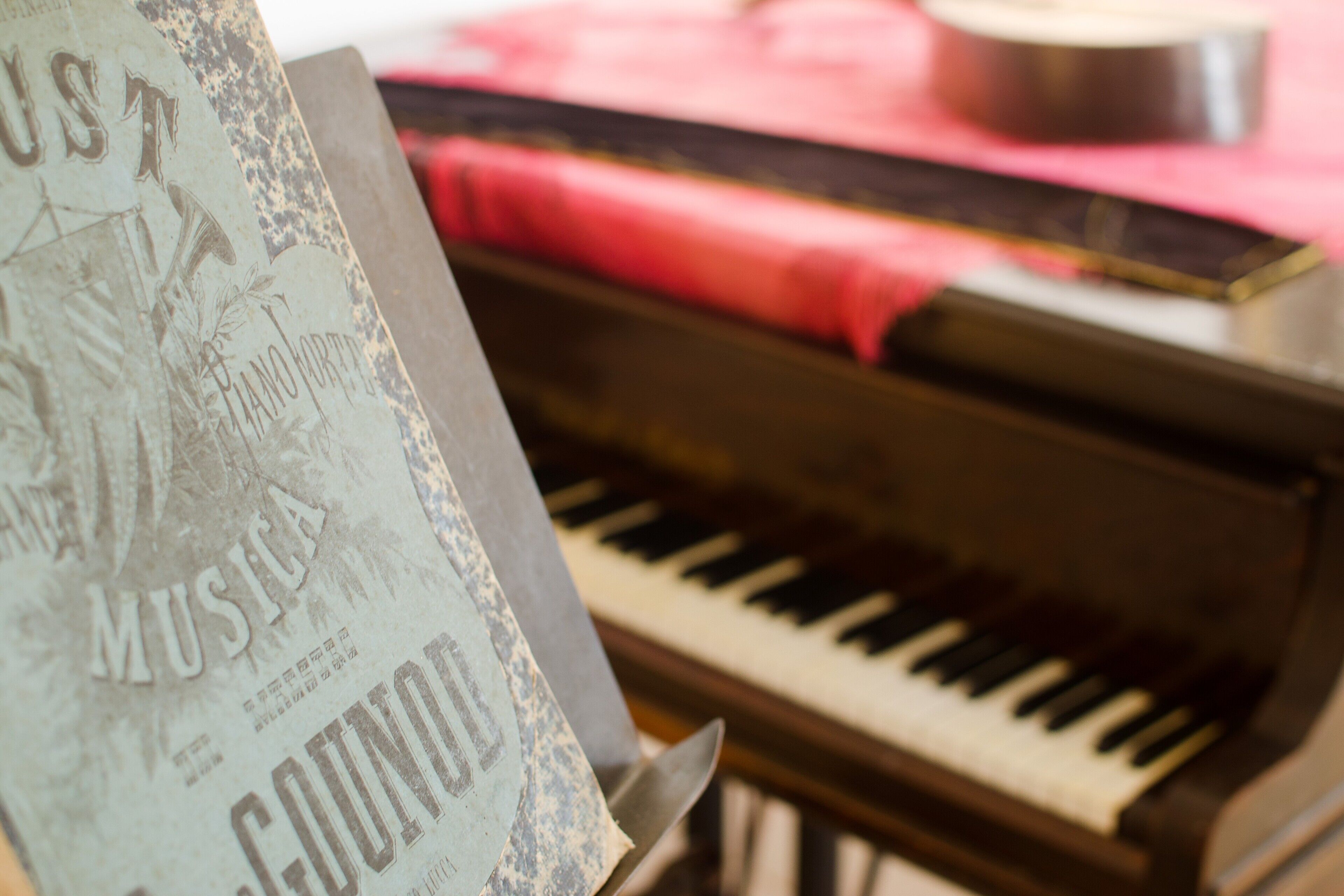 Um piano com um pano vermelho, uma partitura de música intitulada 'MUSICA COUNCIL' e um objeto metálico ao fundo.
