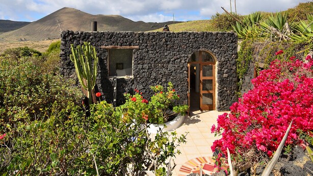 A stone building with a door and windows, surrounded by greenery and a mountainous backdrop. - La Bodega – On a Volcano with Piano, Sea Views, Private Terrace & Wi-Fi (Haria)