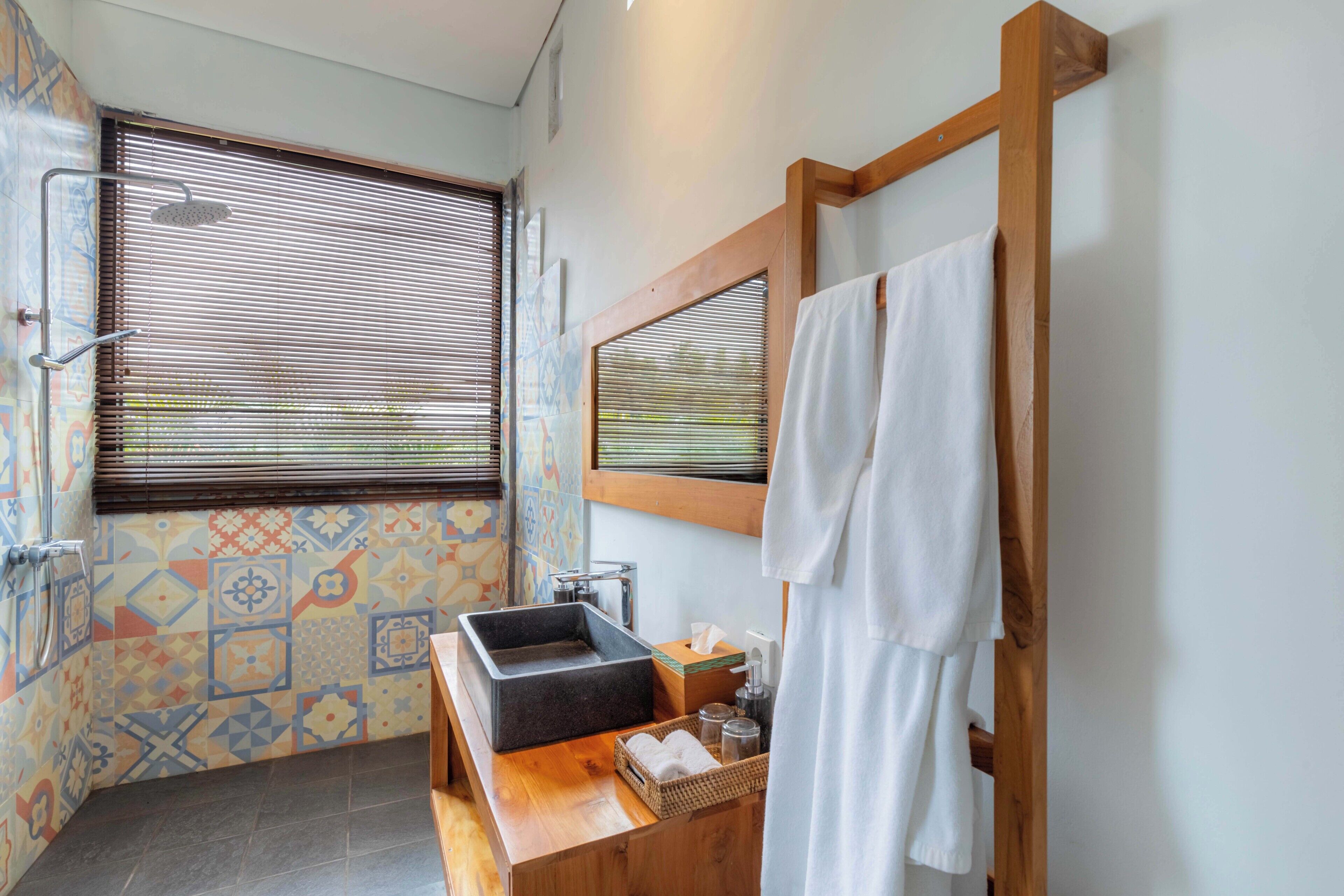 A bathroom with a wooden vanity, a large window with blinds, a shower, and a tiled wall.