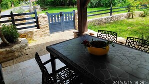 A covered patio with a table and chairs, a yellow bowl with a flower, and a view of a green garden and a blue gate. - Stone country house (Saint-Remy)
