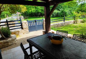 A covered patio with a table and chairs, a yellow bowl with a flower, and a view of a green garden and a blue gate.