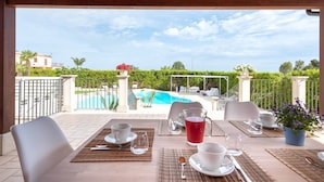 An outdoor dining area with a view of a swimming pool, surrounded by greenery and a wooden pergola. - Modica Country Villa - Villa with pool (Modica)