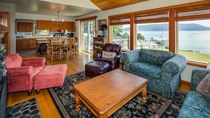 Un vaste espace de vie doté d’un plafond en bois, de grandes fenêtres offrant une vue sur la mer, un coin repas avec des meubles en bois et un coin salon confortable.