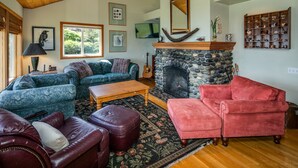 Un salon chaleureux avec une cheminée en pierre, un plafond en bois et divers sièges, notamment un fauteuil rouge et un canapé bleu.