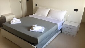 Een netjes opgemaakt bed met witte lakens en een grijze deken, een nachtkastje met een lamp, en een klein dressoir.