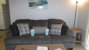 Un salon avec un canapé gris, des coussins à motifs, une table basse avec des magazines et un carnet, du pop-corn dans un récipient rayé et un tableau représentant une vague accroché au mur.