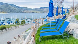 A row of blue lounge chairs with umbrellas, a wooden fence, and a view of a lake with boats in the background. - I'm Your Huckleberry at Lake Chelan Lookout Vacation Rentals (Chelan)