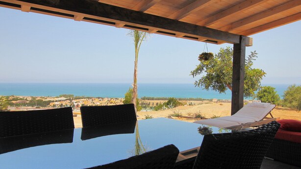 A covered outdoor seating area with a view of the sea and a distant town. - Modern Villa with Private Golf Practice, Pool, Garden, Terrace, Wi-Fi, Sea View (Campofelice di Roccella)