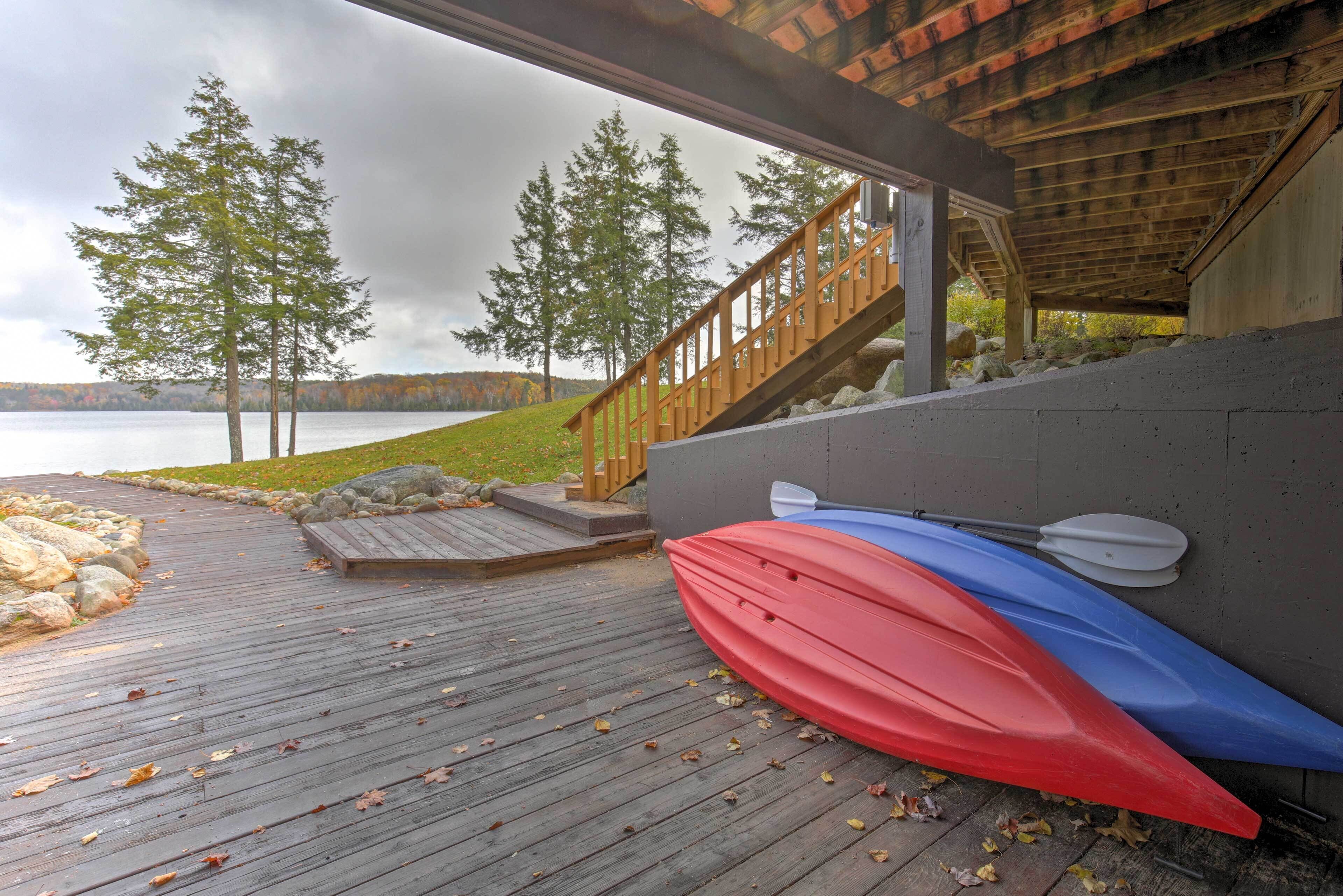 A wooden deck with kayaks and a paddle leaning against a wall, overlooking a lake with trees and a cloudy sky.