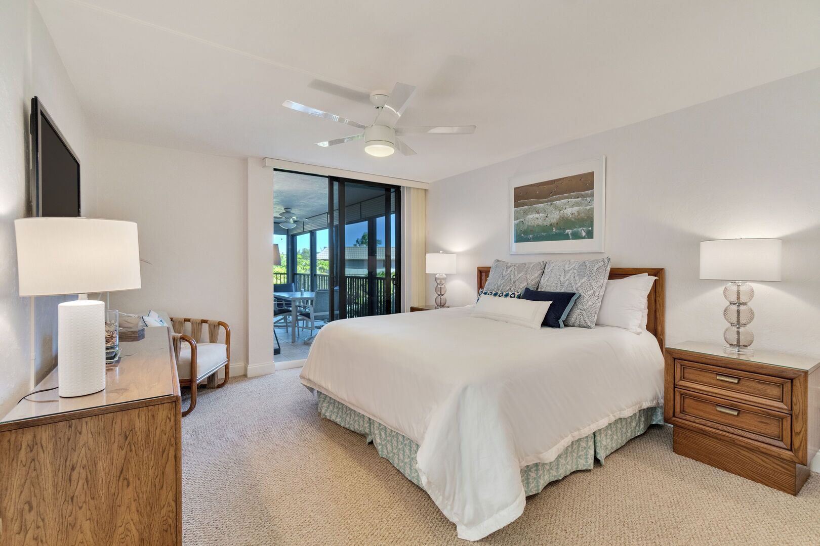 A bedroom with a large bed, a ceiling fan, a nightstand, a desk, and a chair. There is a balcony with a table and chairs outside.