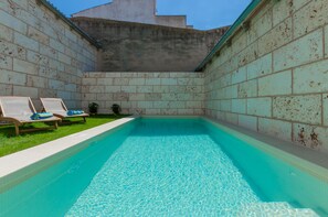 Ein rechteckiger Swimmingpool mit klarem, blauem Wasser, umgeben von einer Ziegelmauer. Auf der linken Seite des Pools befinden sich zwei Liegestühle mit blauen Handtüchern.