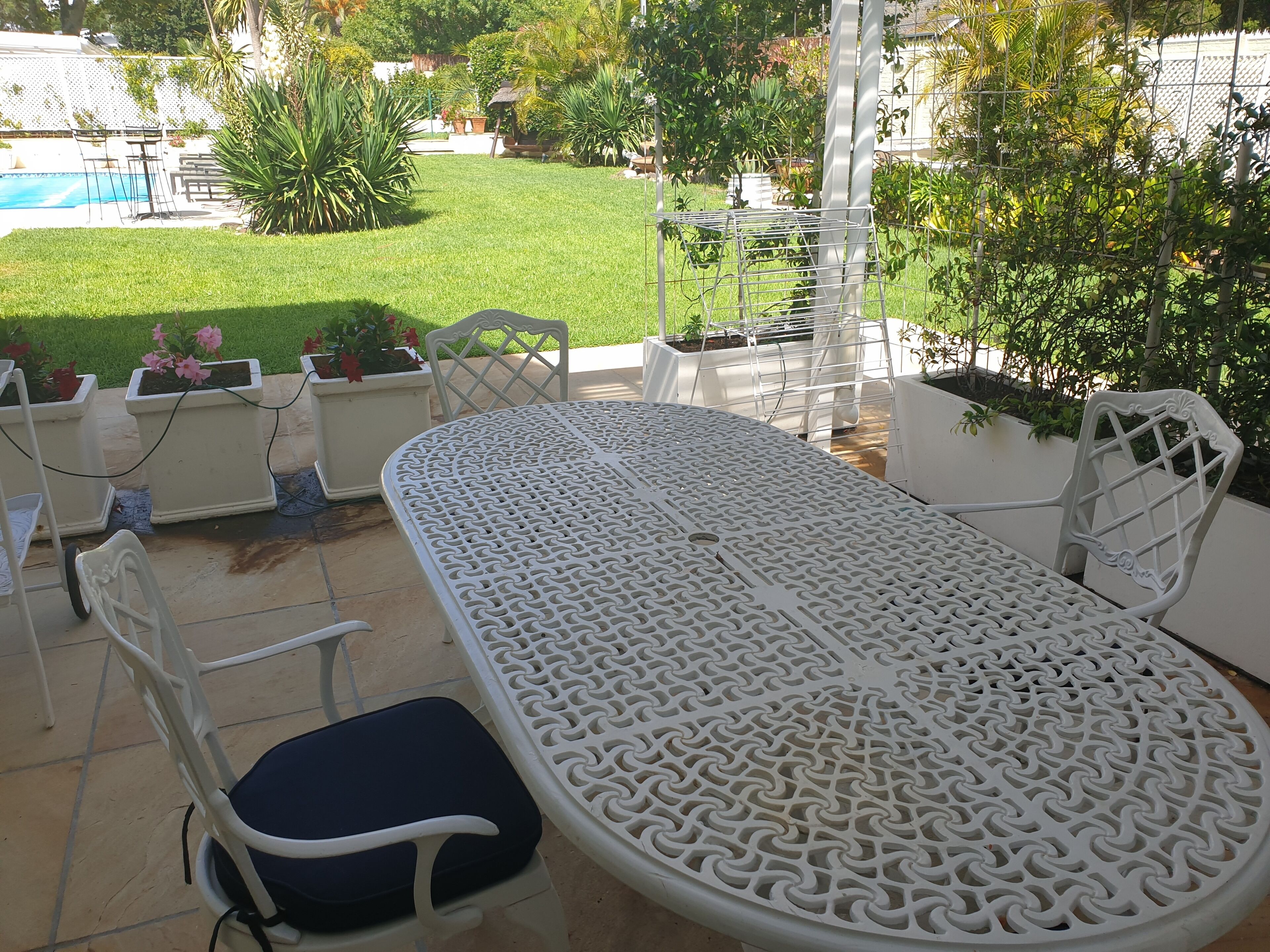 Une terrasse extérieure blanche, avec une table ronde et des chaises, entourée de plantes en pot et offrant une vue sur une piscine.