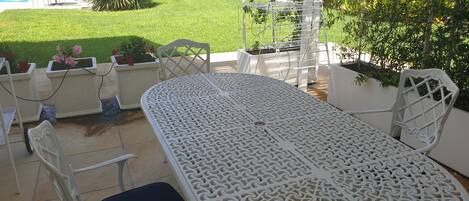 Une terrasse extérieure blanche, avec une table ronde et des chaises, entourée de plantes en pot et offrant une vue sur une piscine.