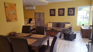 A living room with a sofa, a dining table with chairs, a coffee table, and a window with a view of the outdoors. - 4-STAR-Apartment (AA Superior rated) at the Constantia Wine Route / Kapstad (Constantia)