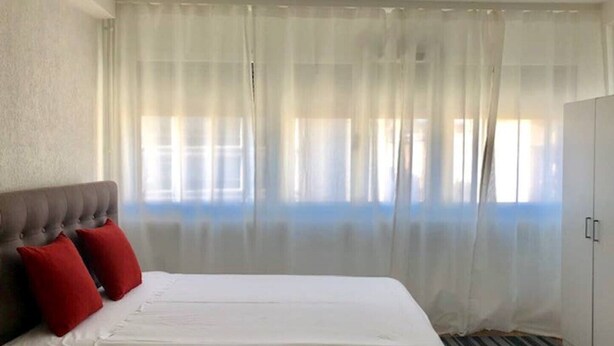 A hotel room with a bed, white curtains, a grey headboard, and a striped carpet. - Rent studio per week (Eaux-Vives)