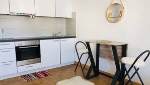 A modern kitchen with white cabinets, a built-in oven, and a black countertop. There is a small round table with two black chairs and a white furry rug on the wooden floor. - Rent studio per week (Eaux-Vives)
