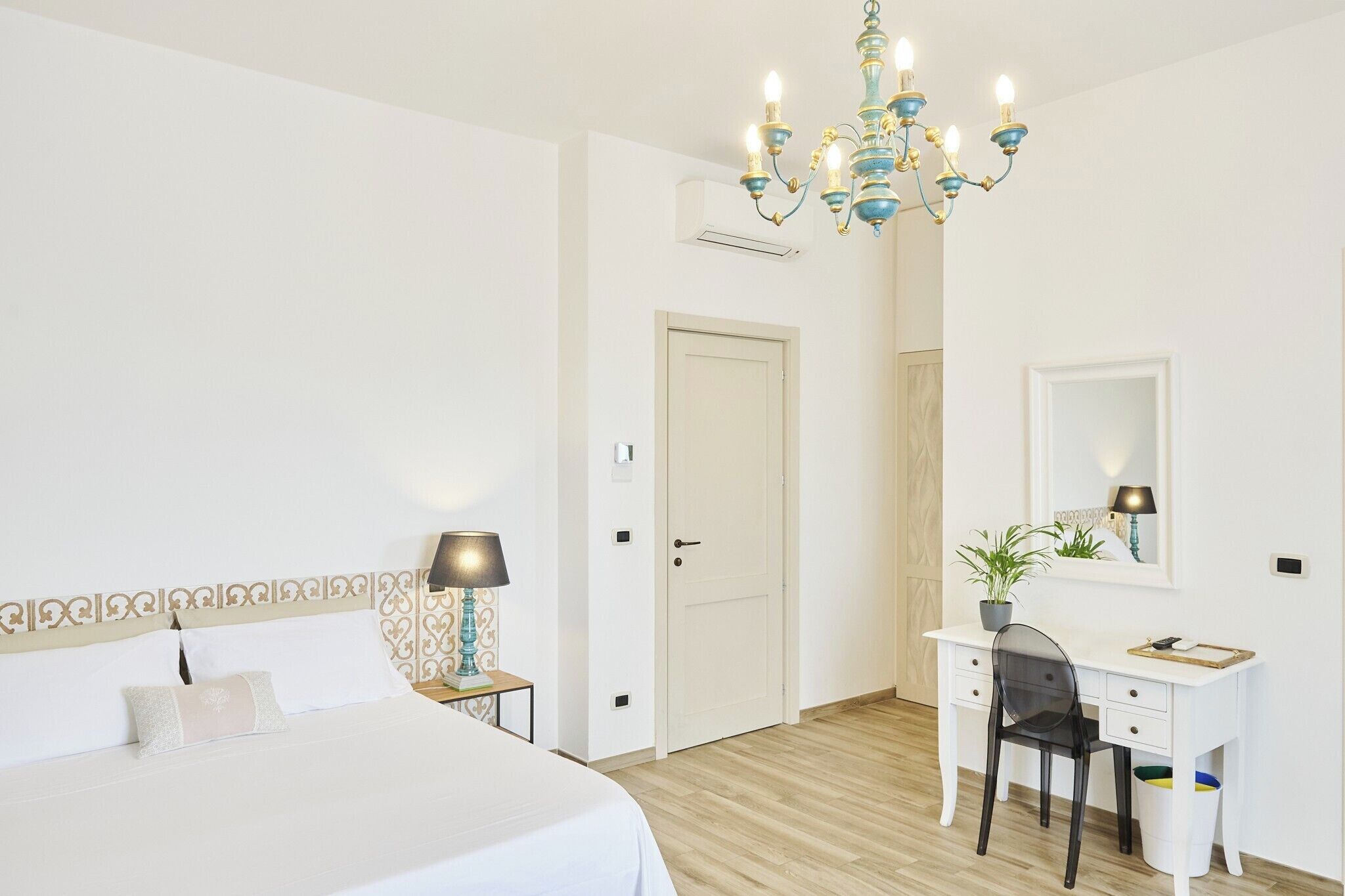 A bedroom with a bed, bedside table, lamp, desk, chair, and a chandelier.