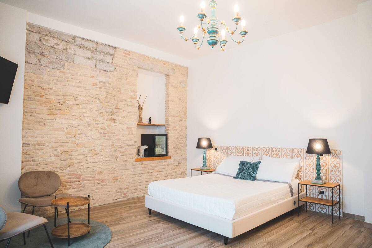 Une chambre avec un mur en briques, un lit, deux tables de chevet avec des lampes, une chaise et un téléviseur posé sur une étagère.