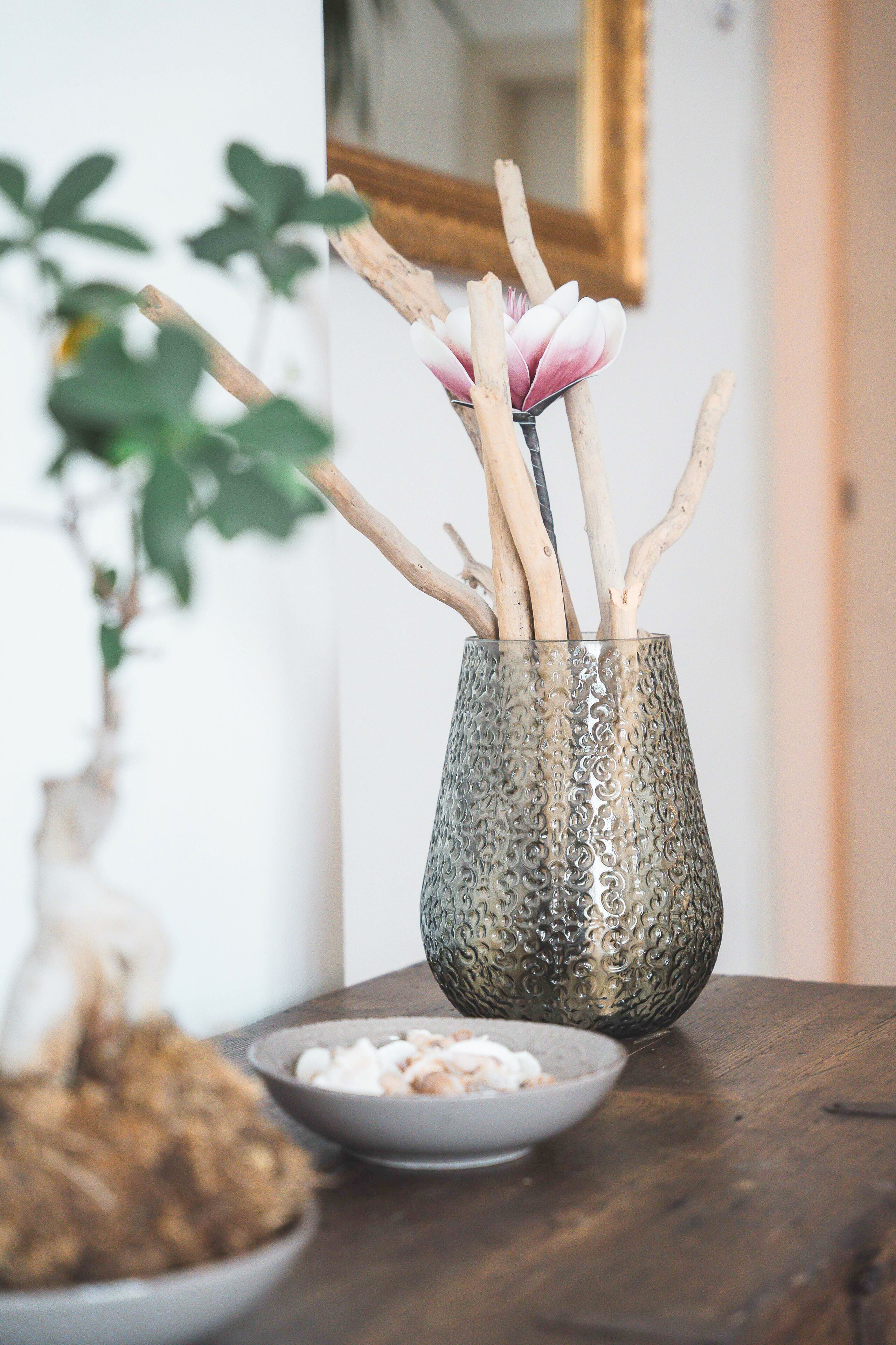 A decorative glass vase with a pink flower and wooden branches on a wooden table.