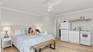 A bedroom with a bed, bedside tables, a bench, a ceiling fan, a kitchen area with a fridge, microwave, stove, and a shelf with dishes.