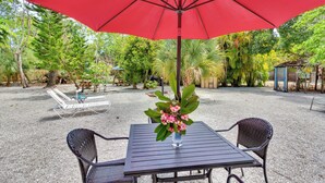 An outdoor seating area with a table and chairs under a red umbrella, surrounded by greenery.