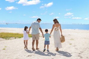 Eine vierköpfige Familie hält Händchen am sandigen Strand, im Hintergrund ein klarer Himmel und ruhiges Meer.
