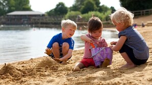 Drie kinderen spelen op een zandstrand, met water en bomen op de achtergrond.