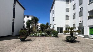 Ein Innenhof mit gefliestem Boden, umgeben von weißen Gebäuden mit zahlreichen Fenstern und Balkonen. Es gibt Kübelpflanzen mit Blumen und Sträuchern.