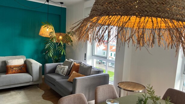 Ein modernes Wohnzimmer mit grauem Sofa, einem Esstisch mit Stühlen, einer hängenden Lampenschirm aus Gewebe und Blick ins Freie durch große Fenster.