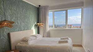 Ein ordentlich bezogenes Bett mit weißen Leinentextilien, Blick auf die Stadt durch große Fenster und eine grüne, strukturierte Wand.