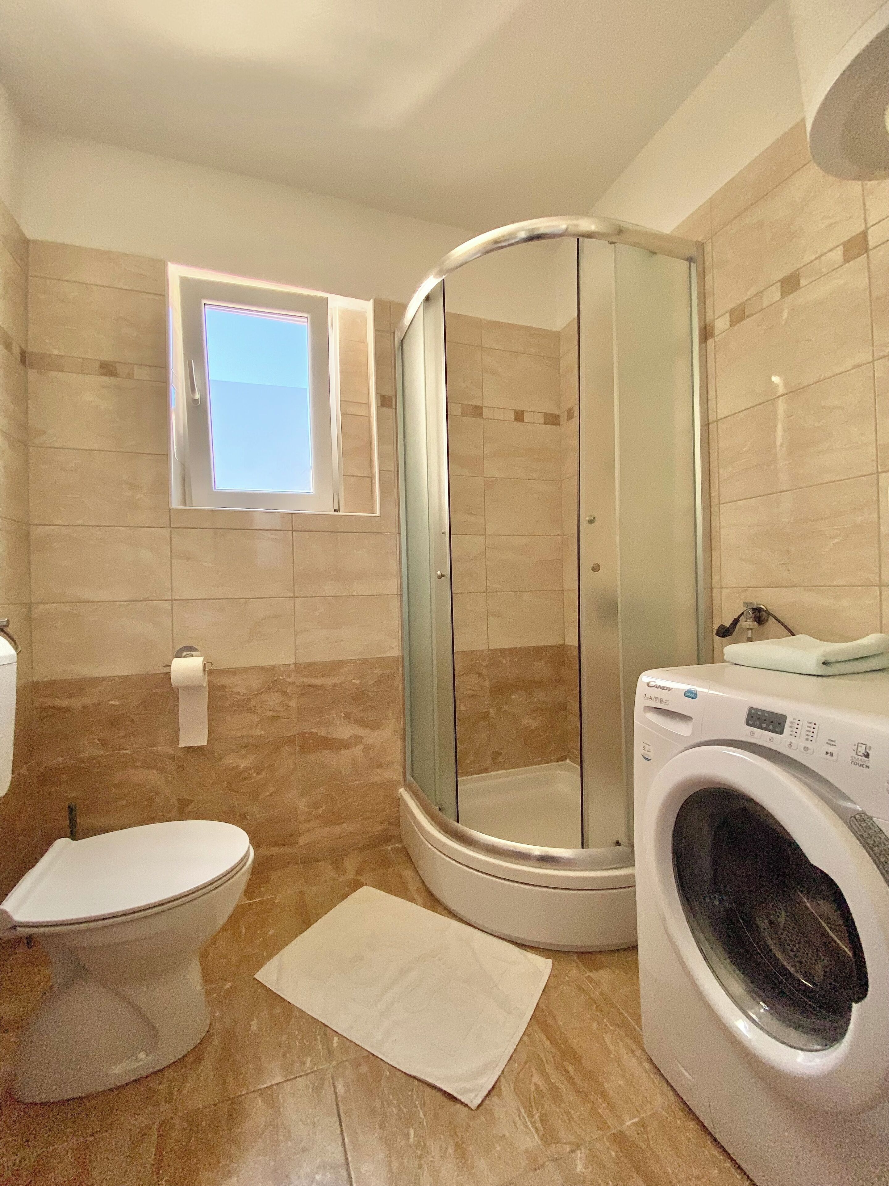 Une salle de bain avec une douche à parois de verre, des toilettes, un lave-linge et une petite fenêtre.