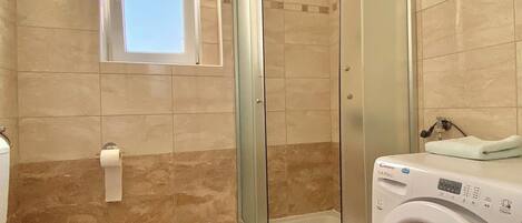 Une salle de bain avec une douche à parois de verre, des toilettes, un lave-linge et une petite fenêtre.