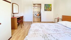 Quarto com cama, escrivaninha, cadeira, pequena mesa com um vaso e um quadro na parede.