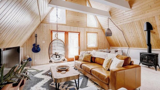 A cozy loft-style room with a sloped ceiling, a leather sofa, a hanging chair, a guitar, and a wood-burning stove. - The Fox A-Frame | An Intimate Mountain Retreat (Blue River)