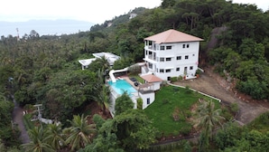 A multi-story building with a swimming pool, surrounded by dense greenery and a hilly landscape.