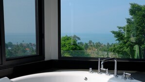 A modern bathroom with a large bathtub, two windows, and a view of the ocean and greenery.