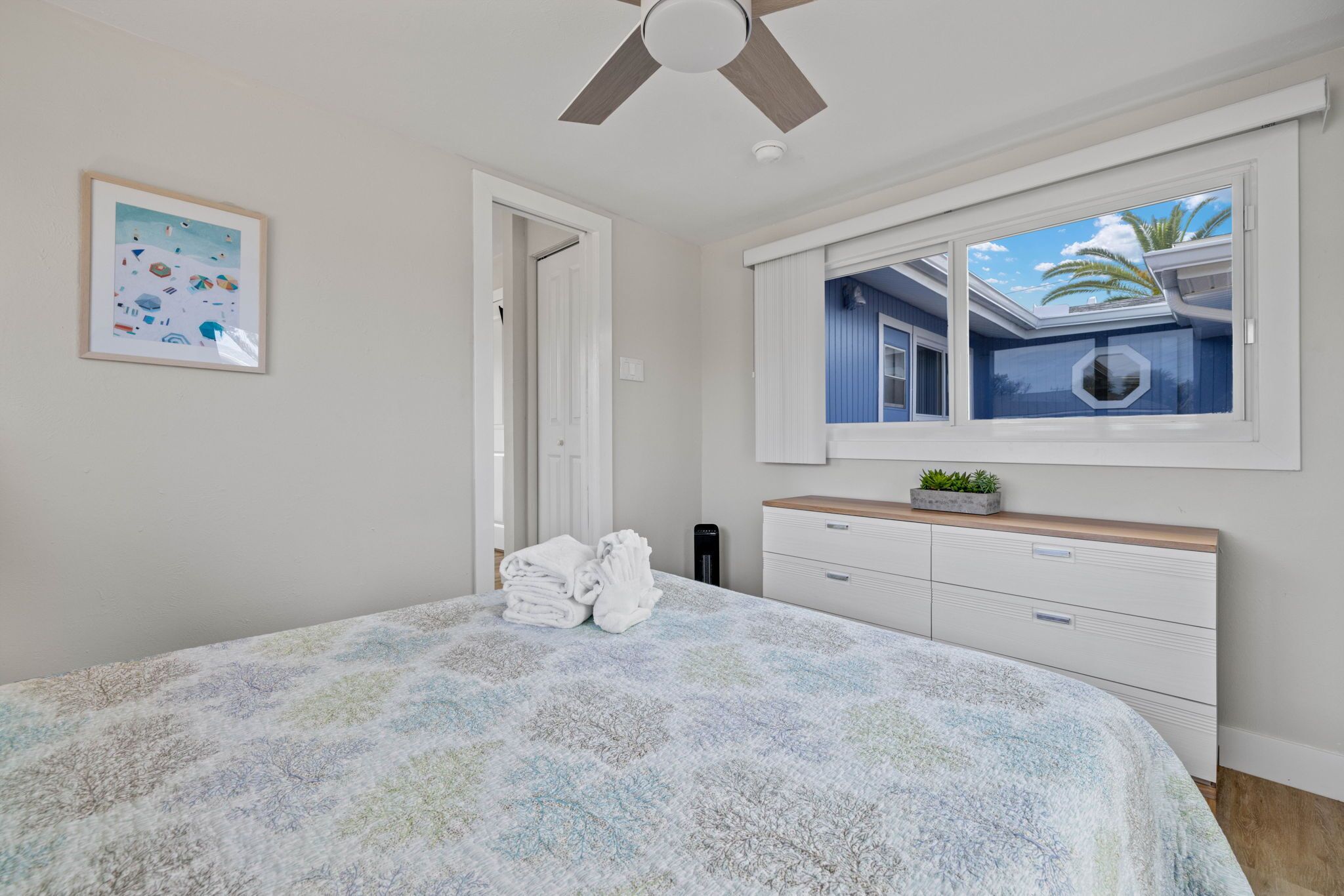 Une chambre à coucher avec un lit, un ventilateur de plafond, une fenêtre donnant sur une maison, un tableau encadré au mur et une commode.