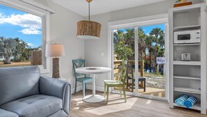 Una sala moderna con un sofá gris, una mesa redonda y dos sillas. Se aprecia una lámpara colgante de mimbre, una estantería blanca con un microondas y vistas a palmeras a través de la puerta de cristal.