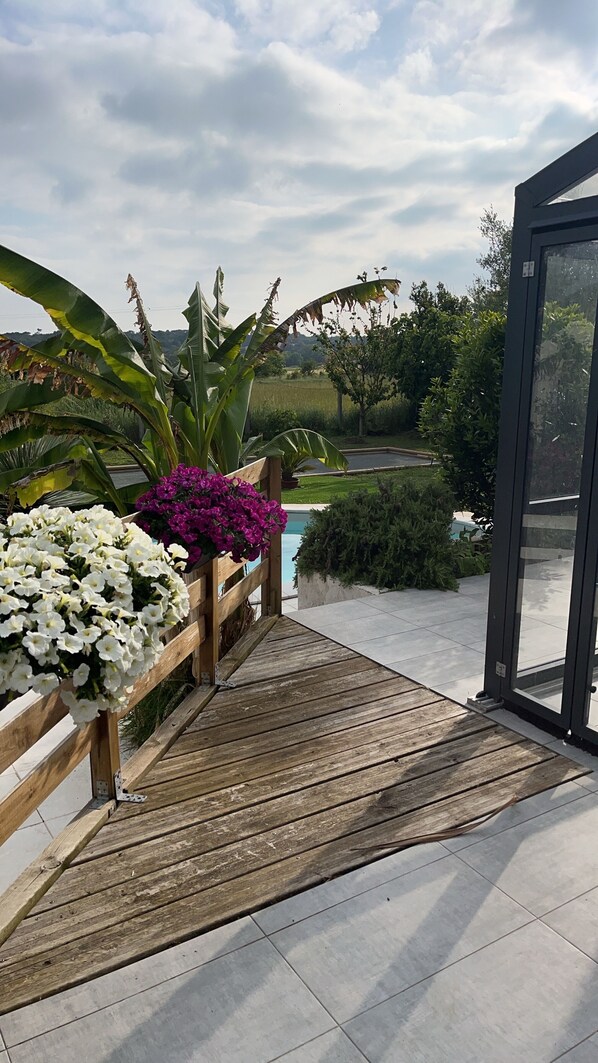 A wooden deck leading to a pool area with white and purple flowers, surrounded by lush greenery and a clear sky.