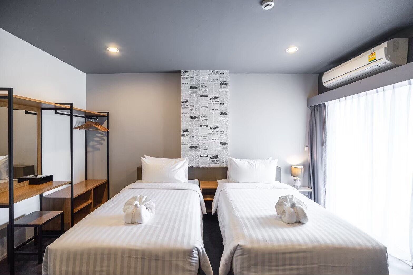 Habitación de hotel con dos camas, un armario de madera, un espejo, un aire acondicionado instalado en la pared y una ventana con cortinas.