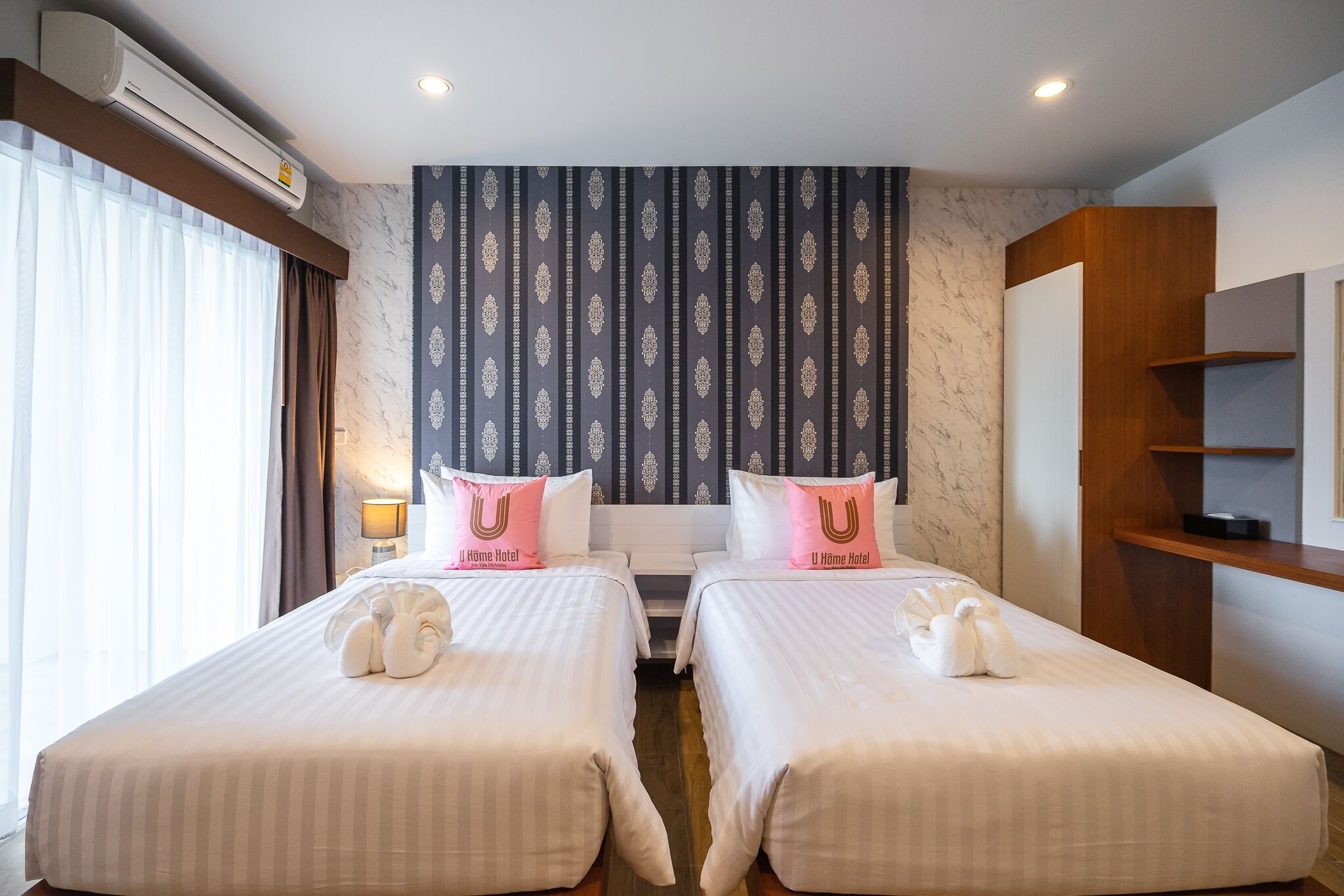 A hotel room with two beds, a large window with curtains, an air conditioning unit, and a patterned wallpaper.