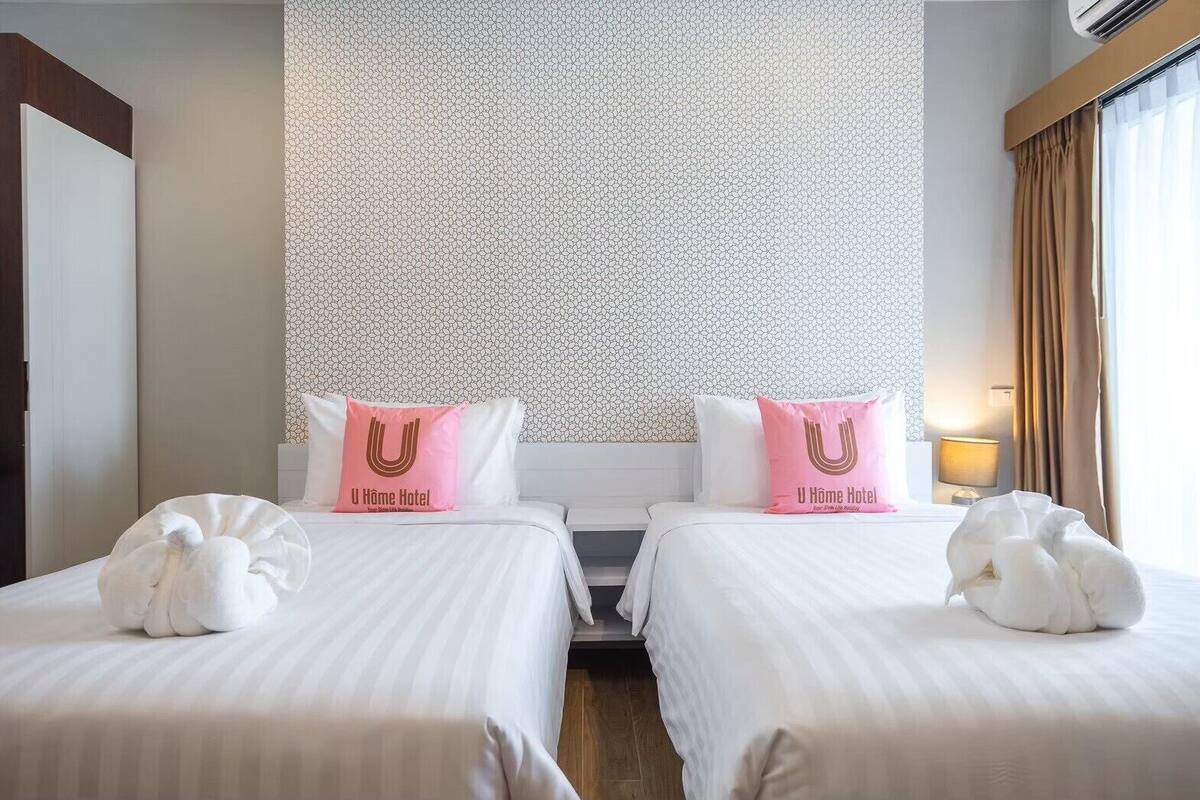 Una habitación de hotel con dos camas, cada una con una colcha blanca y almohadas a juego. La habitación tiene papel tapiz estampado y una ventana con cortinas.