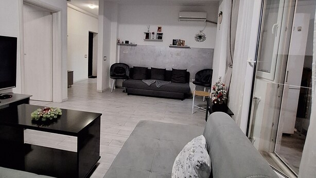 Ein modernes Wohnzimmer mit grauem Sofa, Couchtisch und Fernseher. Ein Glastür führt zu einem Balkon.