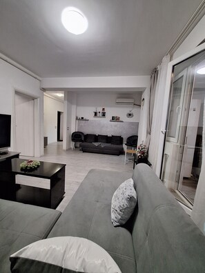 A modern living room with a grey sofa, a coffee table, and a TV. There is a glass door leading to a balcony. - Daniel Residence  Tineretului (Chiajna)