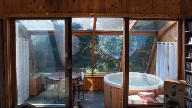 A wooden cabin with a large glass window, a hot tub, a dining table, and chairs.