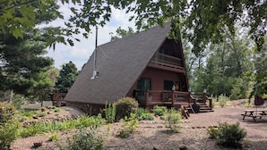 A cabin with a steeply pitched roof, a wooden deck, and a stone pathway leading to a picnic table.